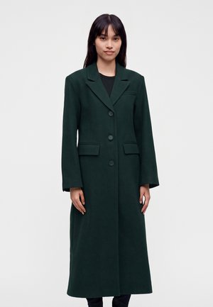 Femme aux longs cheveux foncés portant un long manteau vert foncé boutonné, debout devant un fond clair uni.
