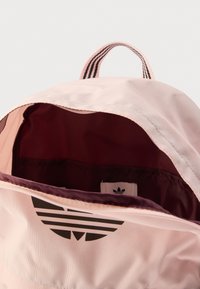 Šviesiai rožinė kuprinė su tekstūruota medžiaga, vidus bordo spalvos, priekyje juodas logotipas su trimis dryžiais. Atviras viršus.