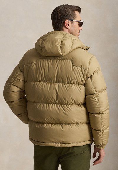 Man wearing beige puffer jacket with hood and dark sunglasses, shown from the back against a plain light background.
