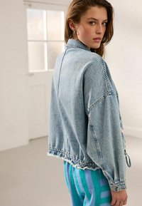 Veste en denim bleu clair avec une coupe ample, longueur cropped, ourlet effiloché et manches décontractées. Portée sur un pantalon à rayures, avec un motif bleu et vert.