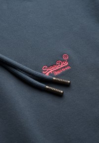 Marineblauwe sweatshirt met een roze geborduurd "SuperDry" logo. Twee donkerblauwe trekkoorden met metalen uiteinden gelabeld "SUPERDRY." Gladde textuur.