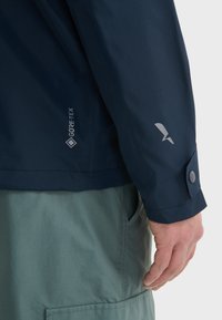 Mornarski GORE-TEX jakna s uskim, vodootpornim dizajnom. Ima elegantnu siluetu, sivi detalj loga i manžete sa dugmadi.