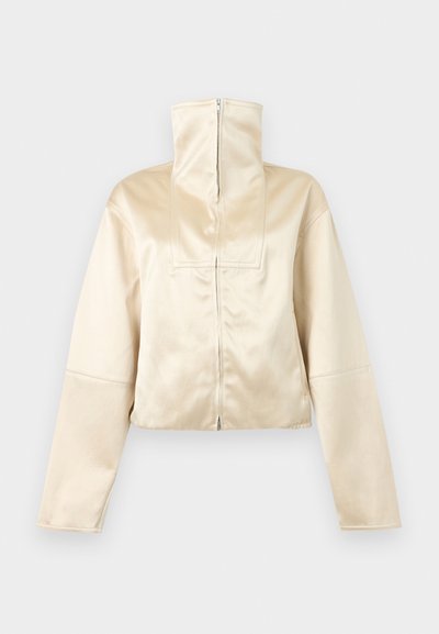 Μπεζ cropped μπουφάν με μακριά μανίκια, ψηλό γιακά με φερμουάρ και κλείσιμο με φερμουάρ μπροστά, εμφανίζεται σε απλό ανοιχτό φόντο.