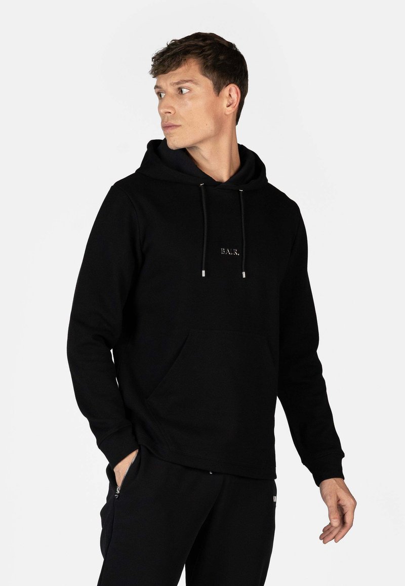 Sudadera negra hecha de una tela suave, con un bolsillo frontal, cordones y un pequeño logotipo bordado. Combinada con pantalones negros.