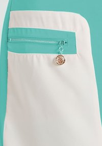 Tejido blanco y turquesa con un bolsillo horizontal de cremallera turquesa que incluye un charm metálico redondo con un diseño de corte floral.