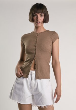 Ung kvinde med kort brunt bobhår, iført en brun knapbluset top og hvide frynset denimshorts, stående mod en ensfarvet baggrund.