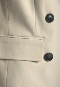 Beżowa kurtka z materiału zbliżeniowym ujęciem czarnego guzika i bocznej kieszeni. Gładka tekstura z widocznymi detalami przeszycia.