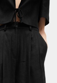 Top cropped en lin noir avec une fermeture à nouer au cou, associé à un pantalon en lin noir taille haute plissé avec poches latérales.