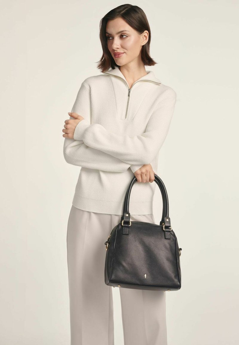 Kobieta w białym swetrze z pół-zipem i jasnych spodniach, trzymająca czarną skórzaną torebkę z podwójnymi uchwytami, patrząca w lewo.