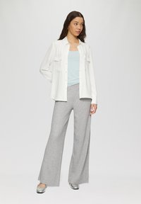 Witte blouse over een lichtblauwe tanktop, gecombineerd met grijsgemêleerde wijde broeken en zilveren platte schoenen. Neutrale achtergrond.