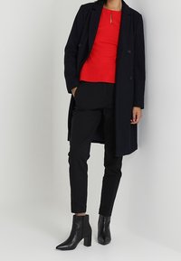 Vrouw in een rode top, zwarte nette broek, zwarte enkellaarsjes en een zwarte lange jas, staande tegen een witte muur.