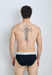Schwarze Baumwollslips mit einem weißen Calvin Klein Logo-Bund. Sichtbares, detailliertes Tattoo eines Kreuzes auf dem Rücken. Natürliche Hauttextur.