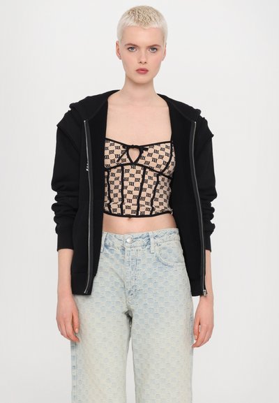 Hanorac negru cu fermoar purtat peste un top scurt bej cu modele geometrice negre, asortat cu blugi largi din denim cu modele în nuanța bleu deschis.