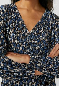 Rypistynyt mekko, jossa on syvä V-aukko ja sinisen, beige ja ruskean kukkakuviot. Pitkät hihat, joissa on ribbitekstuuri.