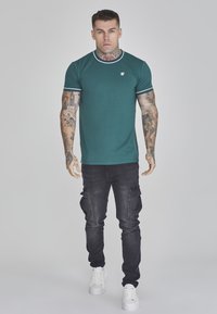 Vyras su tatuiruotėmis, vilkintis žalią marškinėlių, juodus kelnes su kišenėmis ir baltus sportbačius, stovi prieš paprastą pilką foną.