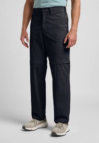 Άνδρας φορώντας μαύρο παντελόνι cargo με φερμουάρ και πλευρική τσέπη, με μπεζ αθλητικά παπούτσια, στέκεται μπροστά από έναν μονόχρωμο γκρίζο φόντο.