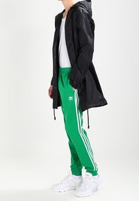 Černá nepromokavá bunda s patentními knoflíky, zelené tepláky s bílými pruhy a bílé sportovní boty s texturovanými detaily.