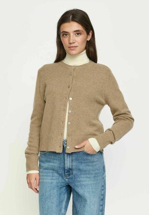 Jeune femme aux longs cheveux bruns portant un cardigan beige à boutons sur un col roulé blanc et un jean bleu, debout avec une main dans la poche.