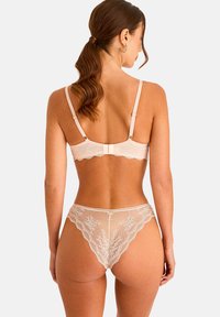 Femme aux cheveux bruns attachés en queue de cheval, portant un soutien-gorge en dentelle rose clair et une culotte en dentelle assortie, vue de dos sur un fond neutre.