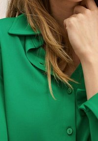 Mujer con cabello castaño claro, vistiendo una blusa verde abotonada con un cuello con volantes, reposando su mano cerca de su mentón.