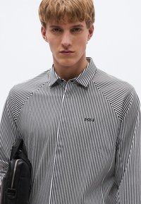Melns un balts svītrains krekls ar pogu apkakli, kreisajā pusē neliels logotips un gluda auduma tekstūra.
