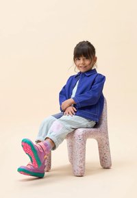 Denim jack in fel blauw, lichtgekleurde overall met patchdetails, en kleurrijke roze en groene boots. Kind zit op een gespikkelde stoel.