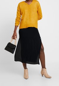 Mosterdgele gebreide trui met V-hals, gecombineerd met een zwarte plissé midi-rok met zijknopen en een hoge split. Beige enkellaarsjes en een kleine zwarte handtas met houten kralenhandvat.