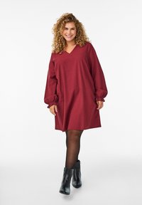 Uśmiechnięta kobieta z kręconymi włosami, ubrana w luźną, długą sukienkę w kolorze burgundowym, czarne rajstopy i czarne botki za kostkę na białym tle.