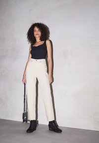 Kvinna med lockigt hår som bär en svart linne, krämfärgade högmidjade byxor och svarta kilklackar, och håller en liten svart handväska.