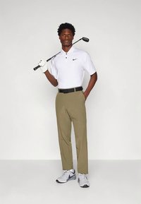 Λευκό πουκάμισο πόλο με γιακά και λογότυπο Nike, καφέ παντελόνι, μαύρη ζώνη και γκρι αθλητικά παπούτσια. Κρατάει μια βέργα γκολφ με ένα λευκό γάντι.