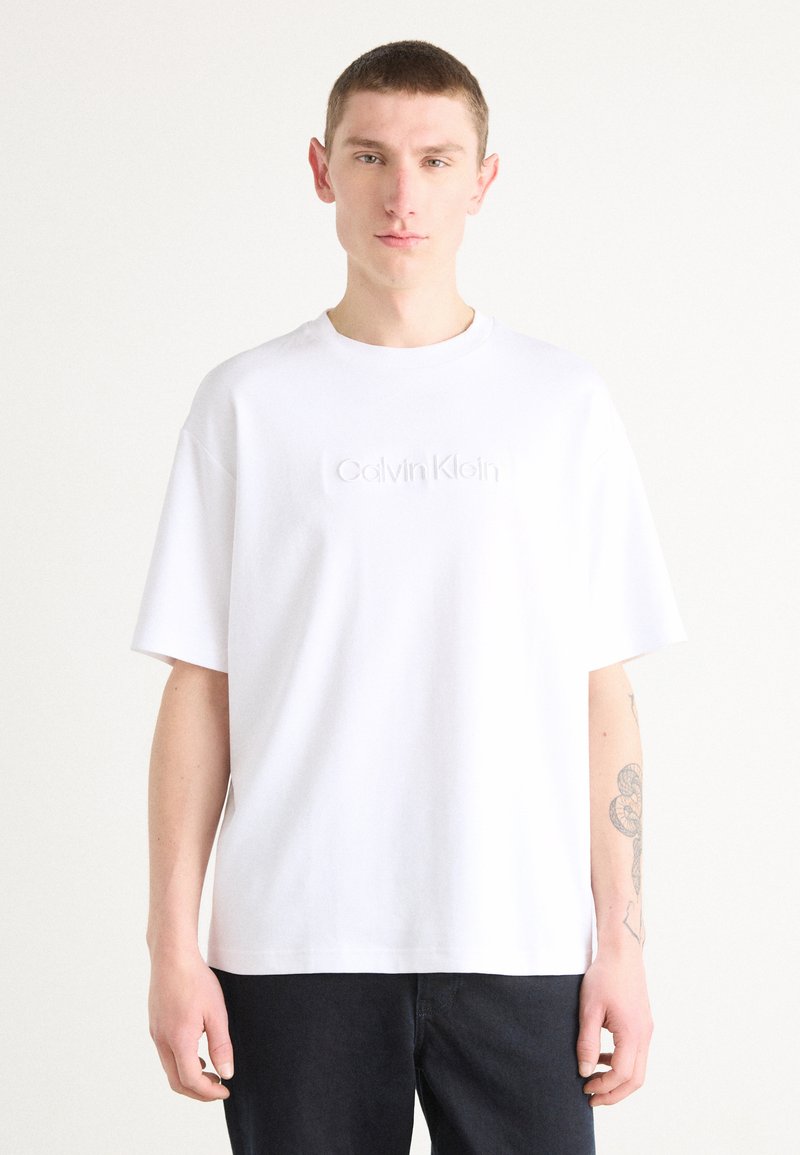 White cotton t-shirt with short sleeves, round neckline, and embossed "Calvin Klein" logo on the front. Smooth texture, relaxed fit.