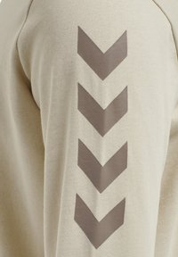 Beiger Stoff mit vier großen dunkelbraunen Chevron-Pfeilen, die nach unten zeigen und vertikal auf dem Rücken angeordnet sind.