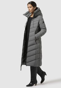 Grijze pufferjas met een lange snit, hoge kraag en grote capuchon. Kenmerkt zich door horizontaal gestikte secties en een ritssluiting.