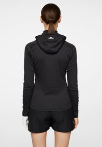 Svart atletisk långärmad tröja med huva, slät textur och logodetalj på baksidan. Kombinerad med svarta shorts och en vit handske.