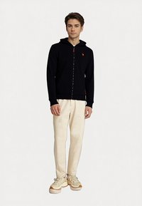 Young man standing wearing black zip-up hoodie, light beige pants, and white sneakers against white background.