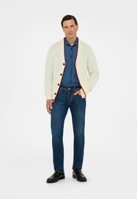 Cárdigan de punto crema con detalles rojos, llevado sobre una camisa de mezclilla azul y jeans azul oscuro, completado con zapatos de cuero negros.