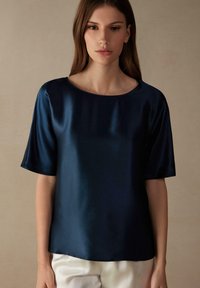Marineblauw satijnen blouse met korte mouwen, ronde halslijn en een gladde textuur, gedragen met lichtgekleurde broeken.