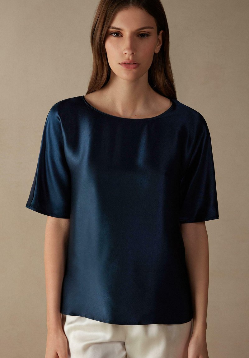 Marineblauw satijnen blouse met korte mouwen, ronde halslijn en een gladde textuur, gedragen met lichtgekleurde broeken.