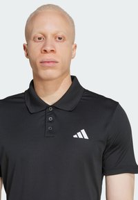 Sort polo skjorte med krave, knap-lukning foran og tre knapper. Har et hvidt Adidas-logo på venstre brystområde. Korte ærmer.