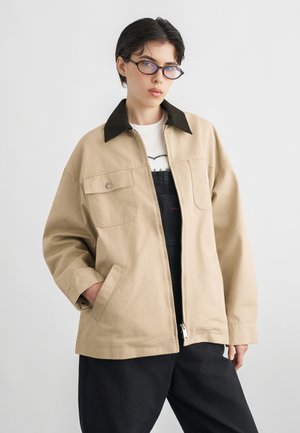 Persoană cu păr scurt și închis la culoare și ochelari, purtând o jachetă bej cu guler negru peste o cămașă albă și pantaloni negri, stând pe un fundal alb.