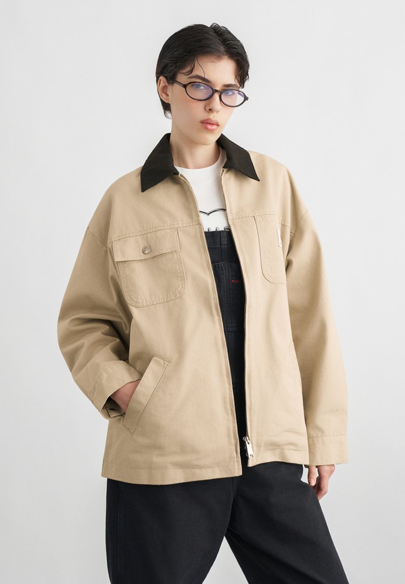 Άτομο με κοντά σκουρόχρωμα μαλλιά και γυαλιά, φοράει μπεζ μπουφάν με μαύρο γιακά πάνω από λευκό πουκάμισο και μαύρο παντελόνι, στέκεται μπροστά σε λευκό φόντο.