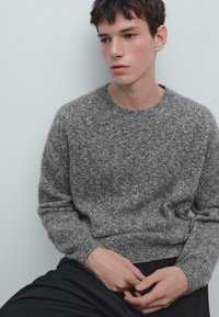 Maglione grigio a maglia con scollo rotondo e polsini a coste. Texture morbida, vestibilità leggermente ampia. Modello seduto, indossa pantaloni neri.