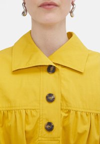Camicetta gialla con colletto, in tessuto testurizzato, dotata di tre bottoni scuri e dettagli arricciati lungo la vita.