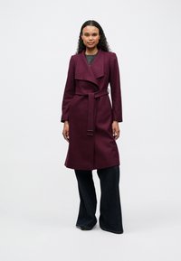Burgundy trenchcoat med bred krage, bälte i midjan och långa ärmar, kombinerat med utsvängda mörka denimbyxor. Slät tygtextur.