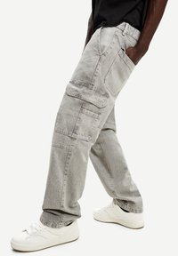 Osoba u svijetlosivim cargo hlačama s više velikih džepova i bijelim tenisicama, stoji s jednom rukom u džepu.