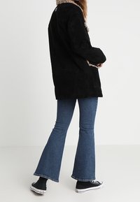 Svart mockajacka med en ljus pälskrage, långa ärmar och sidofickor, i kombination med utsvängda blå jeans och svarta sneakers.