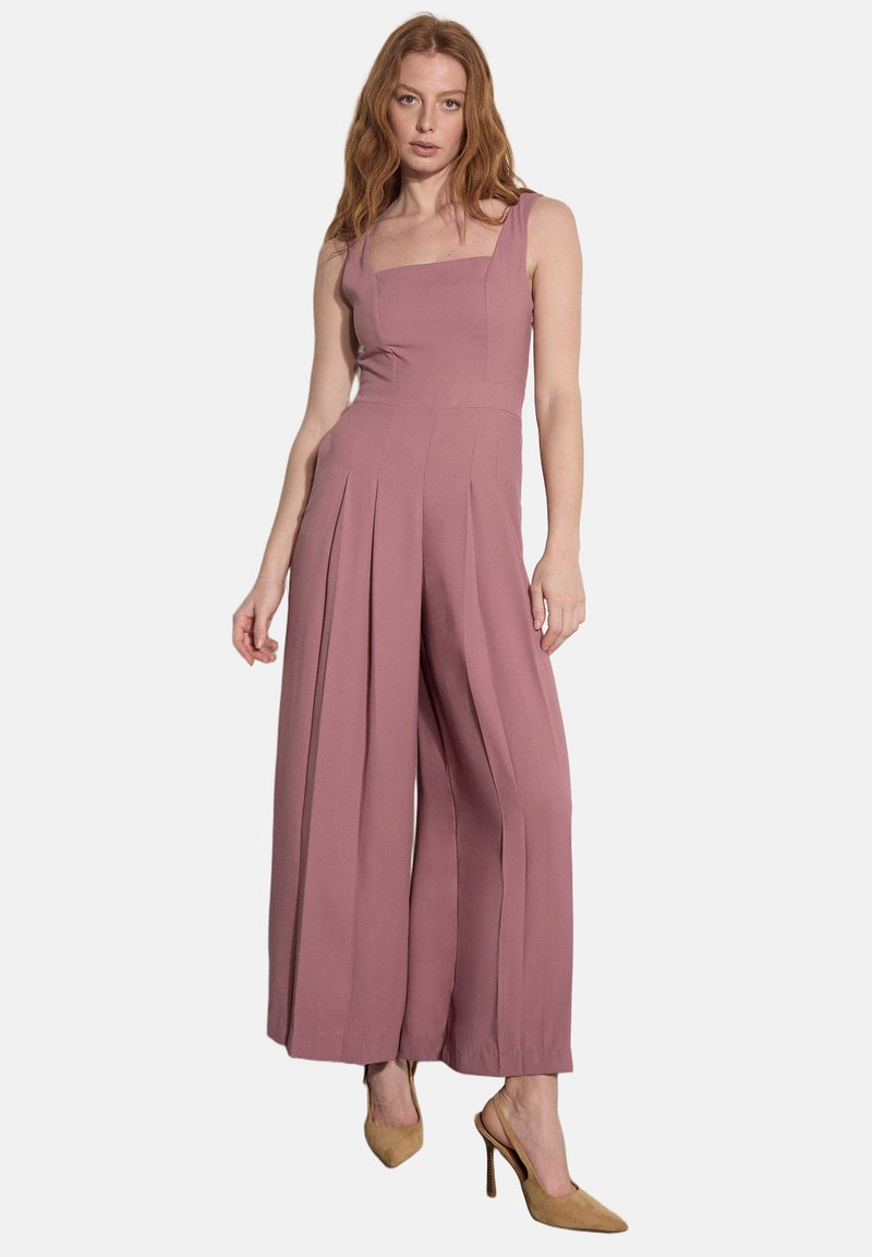 Femme aux longs cheveux roux portant une combinaison sans manches mauve à jambes larges et des escarpins slingback beiges à talons hauts, debout devant un fond blanc.
