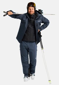 Heren ski-outfit met een marineblauwe waterdichte jas en bijpassende broek, gelaagd met een zwarte gewatteerde vest, terwijl hij witte skistokken vasthoudt.