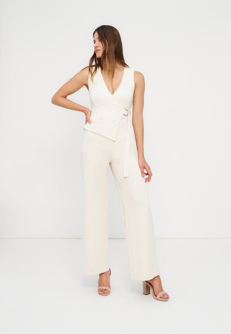 Γυναίκα που φορεί αμάνικο λευκό τοπ με ζώνη και ταιριαστό παντελόνι φαρδύ, ποζάροντας με το ένα χέρι στη μέση και κοιτώντας πλάγια.