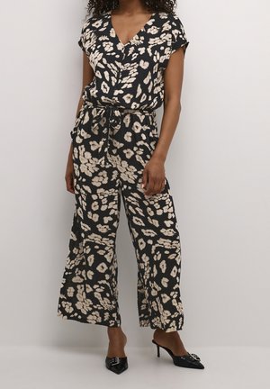 Kobieta ubrana w czarny i beżowy kombinezon z szerokimi nogawkami o kwiatowym wzorze, z krótkimi rękawami, dekoltem w serek i wiązanym w talii, w połączeniu z czarnymi mulesami na obcasie.